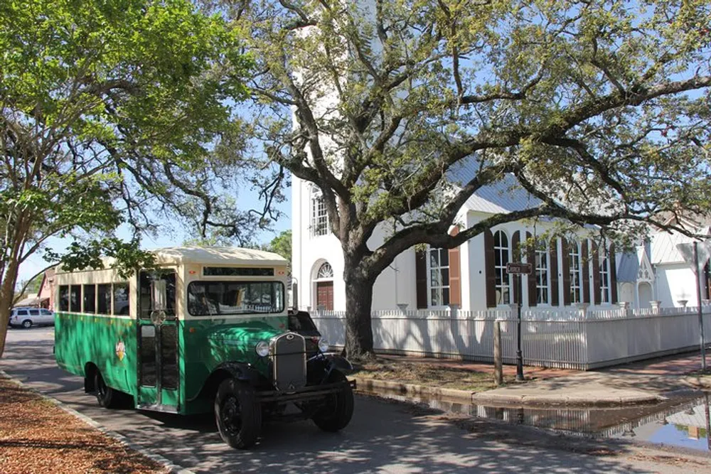 A vintage green and cream bus is parked on a street next to a white building with large windows and a strong presence of greenery under a clear sky