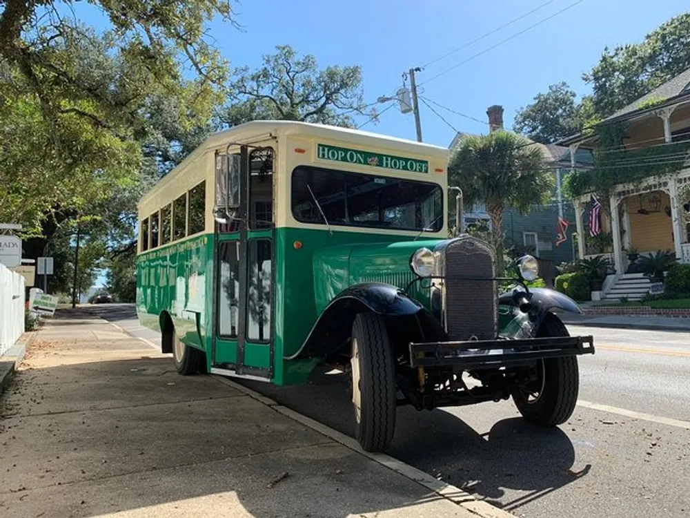 A vintage green and white hop-on hop-off tour bus is parked on a tree-lined street with historical houses in the background