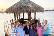 A group of people is smiling for a photo in front of a thatched-roof tiki bar by the water at sunset, with a sailboat visible in the background.
