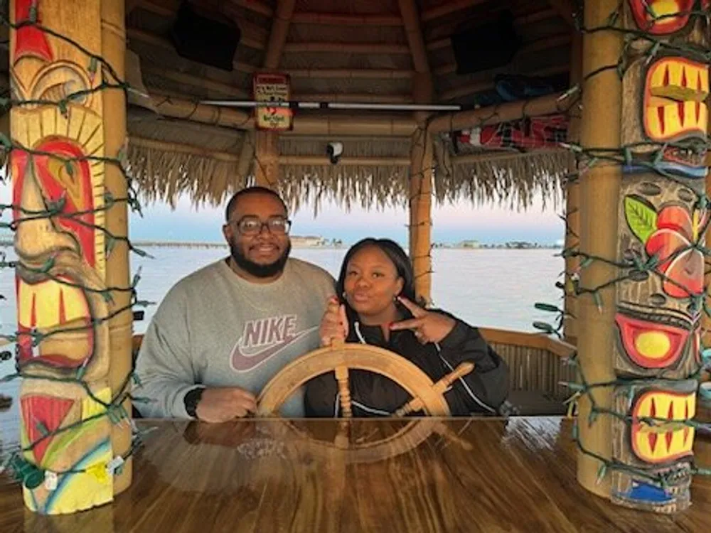A man and a woman pose together behind a wooden wheel likely a decorative ships helm in a tiki-themed setting with a waterfront view in the background