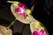Two elegant cocktails garnished with orchids and slices of cucumber are presented on a dark surface, creating a sophisticated and visually pleasing aesthetic.