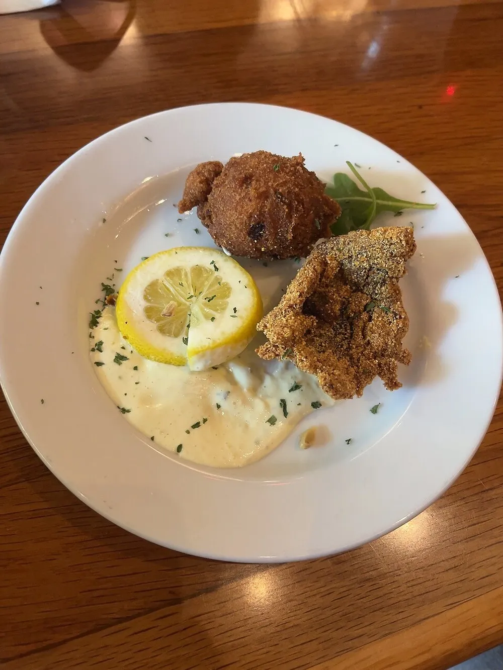 The image shows a plate of food with two pieces of breaded fish a slice of lemon and a dollop of tartar sauce garnished with herbs on a wooden table