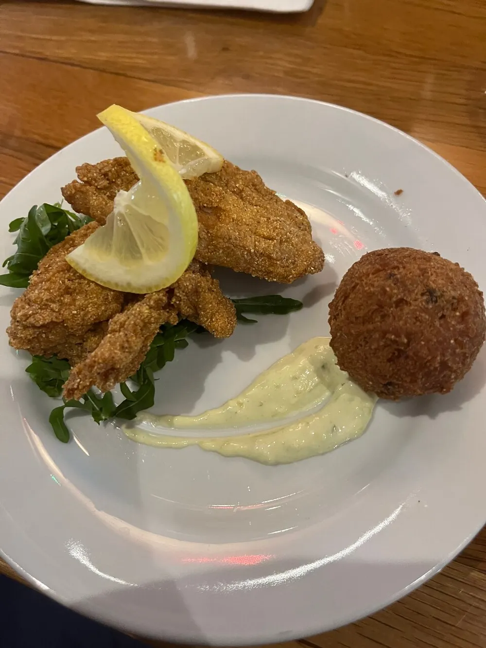 The image depicts a plate of breaded fried food possibly fish garnished with a lemon slice accompanied by a round fried ball and a dollop of creamy sauce on a bed of greens