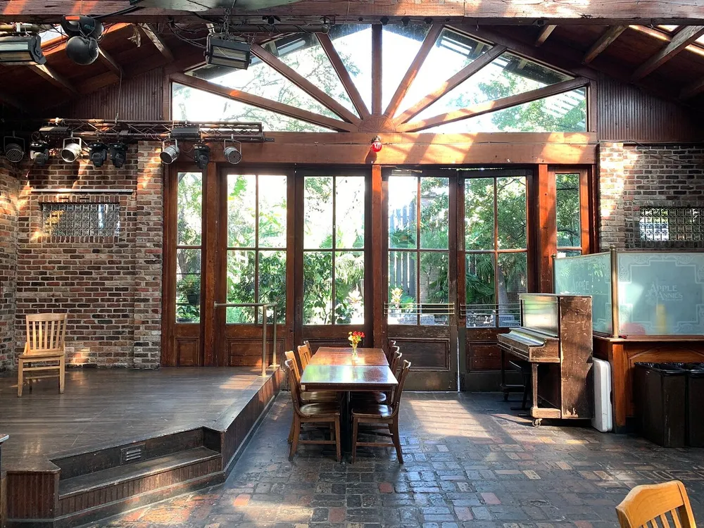 The image shows a cozy rustic room with brick walls wooden beams a stage with lighting equipment and large windows allowing natural light to enter