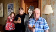 Three people are posing humorously with toy guns and a teddy bear, with a cutout of a classic movie actor in the background, creating a playful scene in a room with vintage decor.