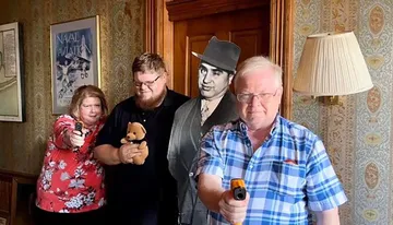 Three people are posing humorously with toy guns and a teddy bear, with a cutout of a classic movie actor in the background, creating a playful scene in a room with vintage decor.