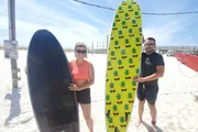 A man and woman are smiling and posing with surfboards on a sunny beach.