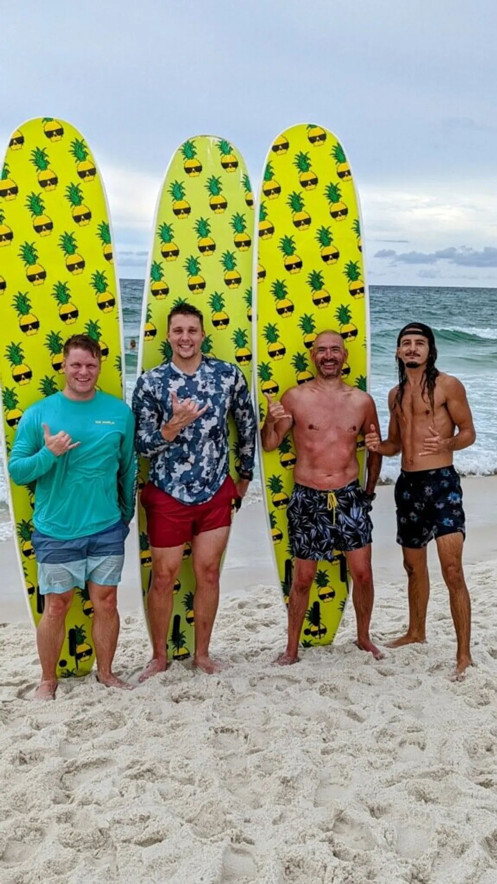 Four individuals are standing on a sandy beach each partially hidden behind bright yellow surfboards adorned with pineapple patterns appearing cheerful with a variety of hand gestures