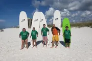 Five individuals are standing on a sunny beach with surfboards, smiling and ready for water activities.