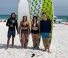 Five individuals are standing on a sunny beach with surfboards smiling and ready for water activities