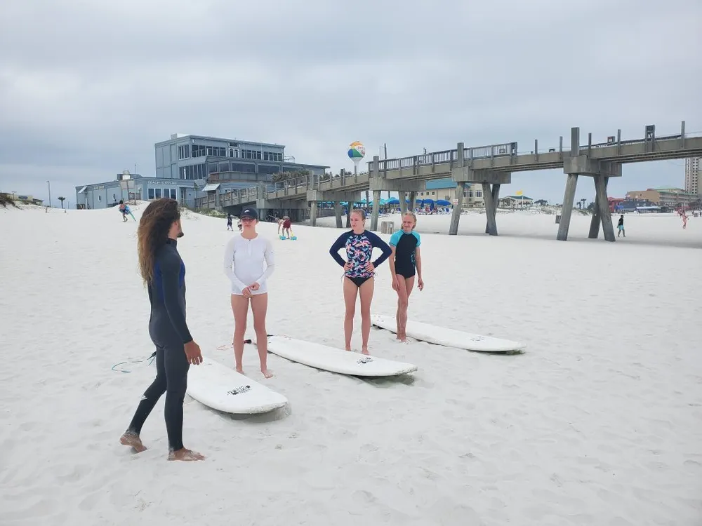 Four individuals with surfboards stand on a sandy beach potentially in a surfing lesson against a backdrop of a pier and beachfront buildings