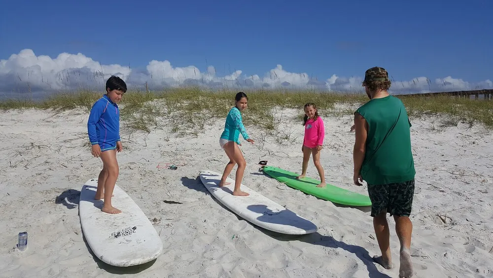 Three children and an adult are standing on surfboards on a sandy beach seemingly preparing for some water activities under a clear blue sky