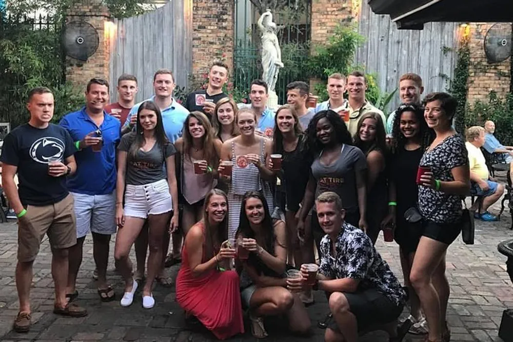 A group of smiling people are posing for a photo in an outdoor setting with many holding drinks suggesting a social gathering