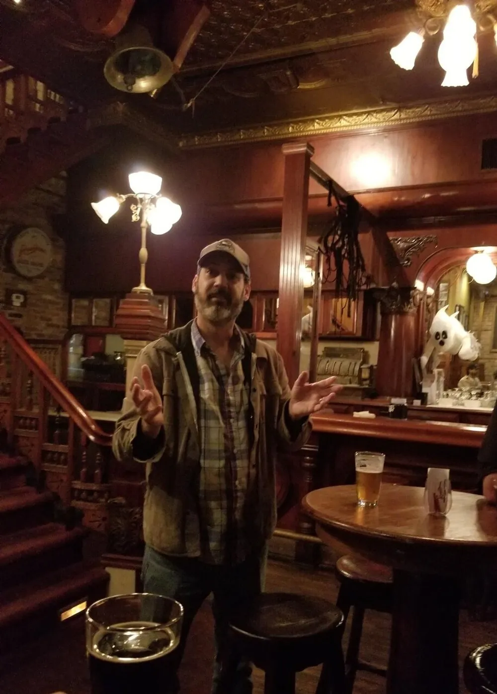 A man in a cap and plaid shirt is standing with his hands open in a dimly lit pub with a beer on the table in front of him