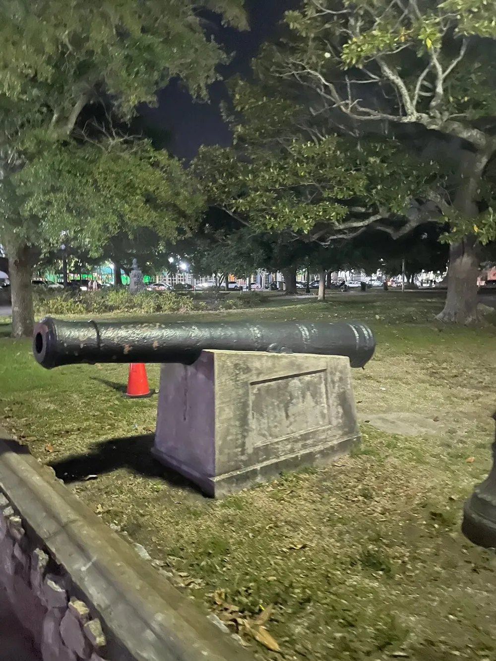An old cannon is mounted on a stone pedestal in a park at night with trees and street lights in the background