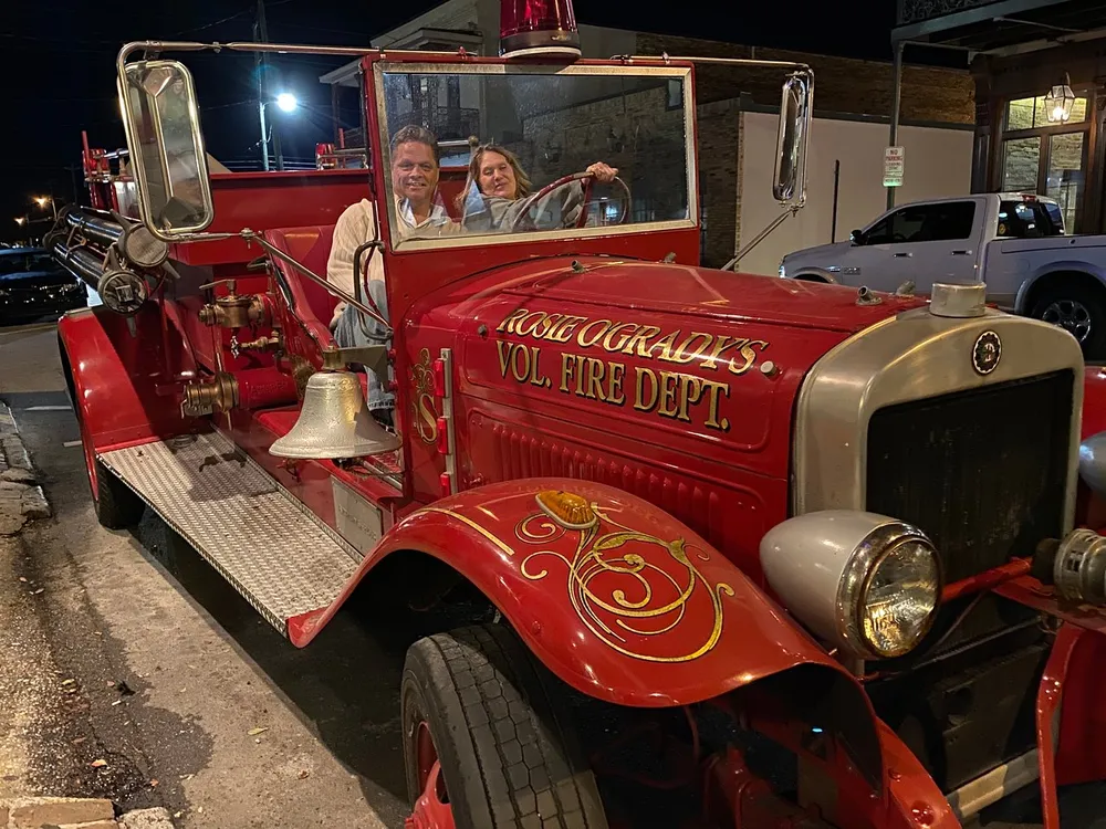 Two people are smiling while sitting on an antique fire engine at night