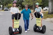 Three people are smiling for the camera, with two women standing on Segways and a man standing between them on a road, all wearing safety helmets.