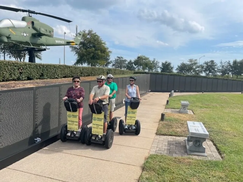 Three individuals are touring an outdoor location on Segways with a military helicopter exhibit in the background
