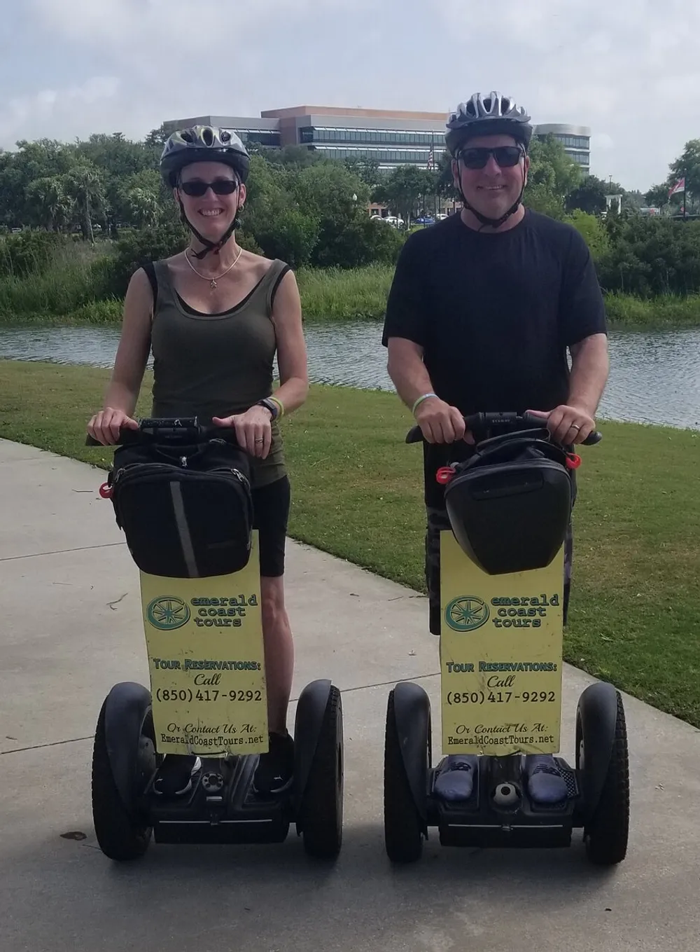 Two people are wearing helmets and smiling while standing on Segway personal transporters in a park-like setting