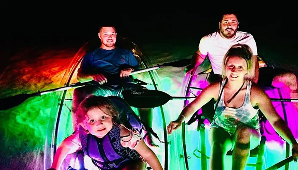 Four individuals including a child are joyfully kayaking at night while their vessel is illuminated with vibrant multicolored lights