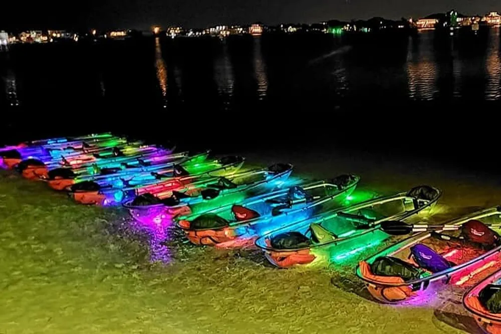 A group of colorful illuminated kayaks is lined up on a beach at night casting vibrant reflections on the water