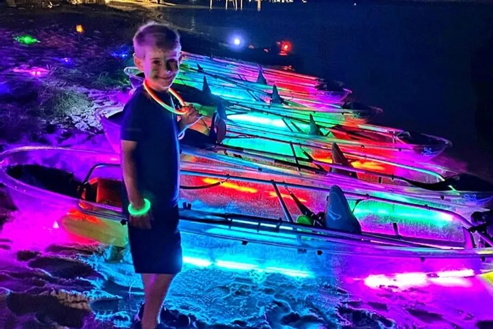 A child is standing on a beach at night with a collection of brightly illuminated colorful kayaks arranged in the background