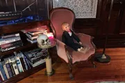 A doll dressed in formal attire is seated on a vintage pink chair beside a small table with a skull and a lit candle, in a room with classic decor and a bookshelf.