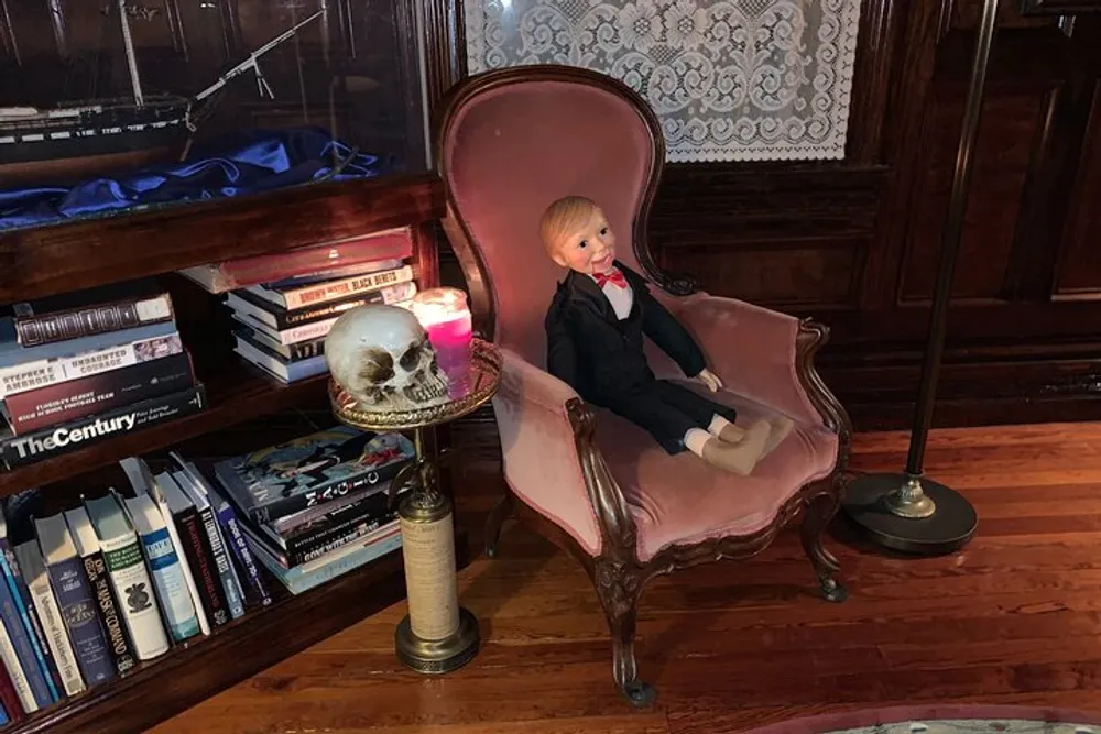 A doll dressed in formal attire is seated on a vintage pink chair beside a small table with a skull and a lit candle in a room with classic decor and a bookshelf