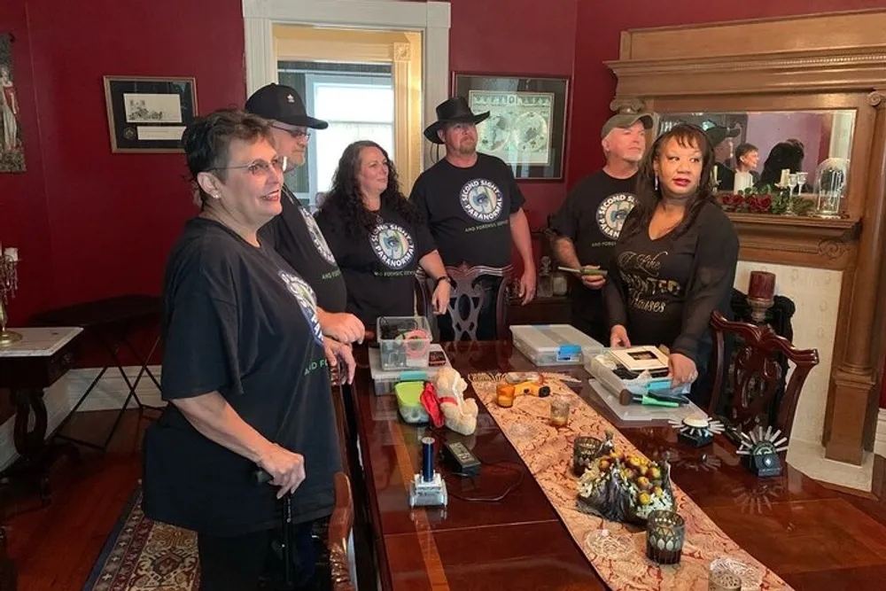 A group of people some wearing coordinated black T-shirts stand in a vintage-style room with various objects on a table in front of them suggesting they could be conducting a gathering or presentation