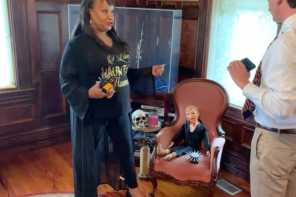 A person is standing in a room with a The Haunted Mansion theme displaying a shirt with the attractions logo next to a vintage armchair with a ventriloquists dummy a skull and other spooky paraphernalia while another person is taking a photo