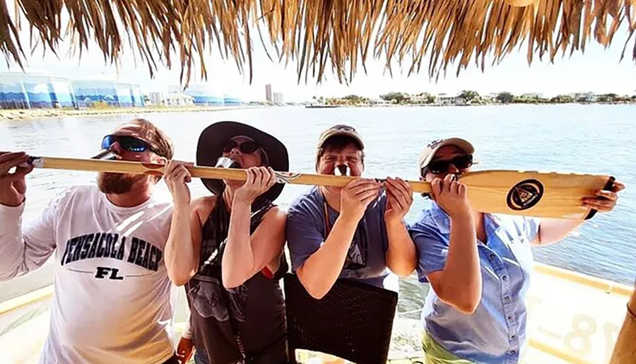 A group of people is playfully biting down on a single oar under a thatched roof, possibly indicating a comical or lighthearted moment during a boating or beach activity.