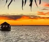 A serene sunset over a body of water with silhouettes of waterfront structures and foliage featuring a gazebo-like boat with people on board