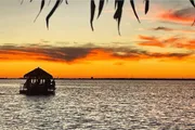 A serene sunset over a body of water with silhouettes of waterfront structures and foliage, featuring a gazebo-like boat with people on board.
