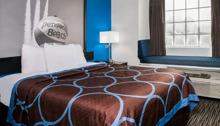 The image shows a well-kept hotel room with a bed that has a brown and blue patterned bedspread a blue accent wall and the words Pensacola Beach on a decorative pillow