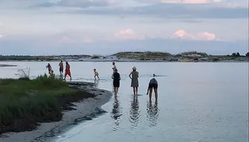 A group of people, including children and adults, are walking along a narrow strip of land that stretches into the water, reflecting the cloudy sky above at dusk.