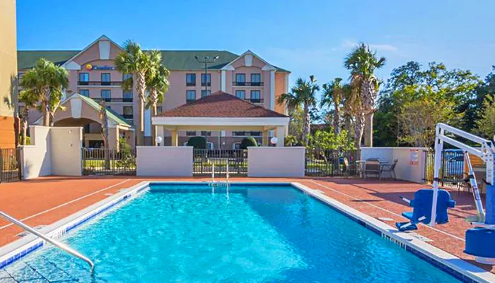 The image shows a sunny outdoor scene featuring a hotel with an inviting swimming pool flanked by palm trees
