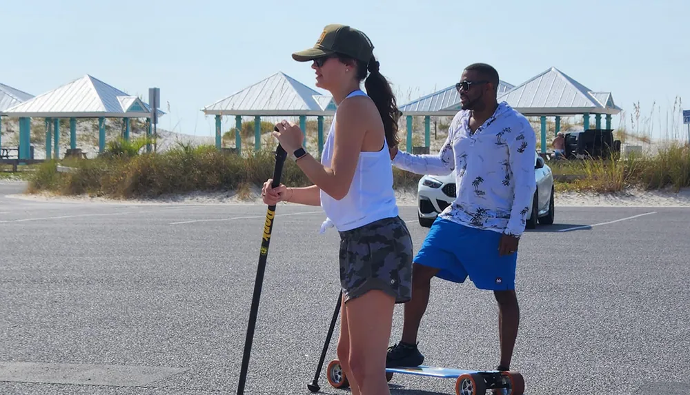 A woman and a man are using what appears to be a two-person land paddleboard on a parking area near the beach