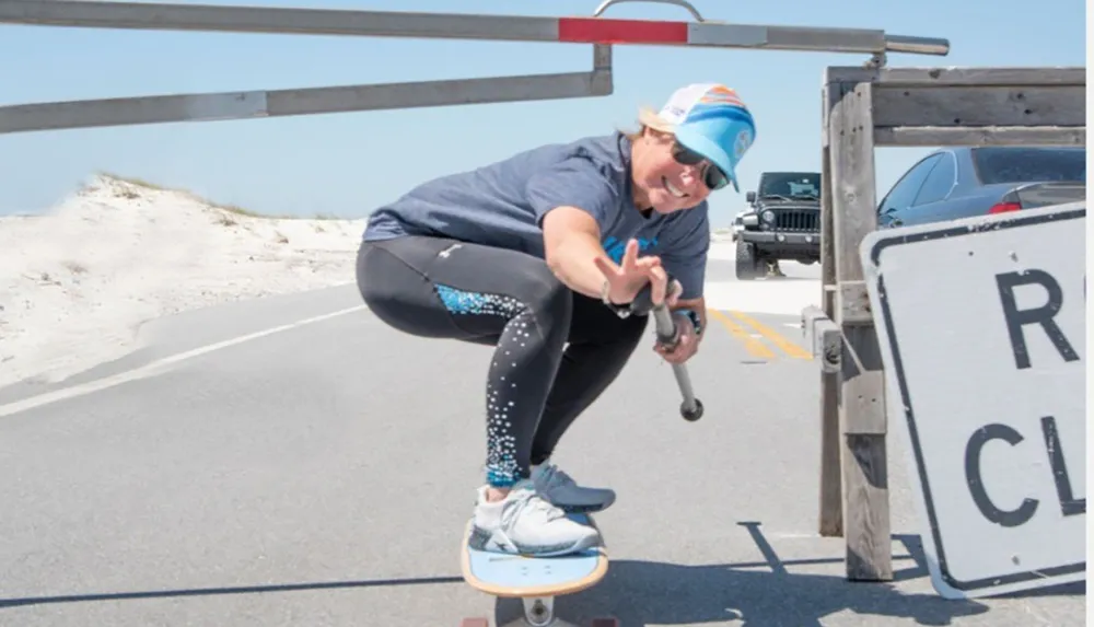 A person is crouching on a skateboard while filming their maneuver with a camera on a sunny day near a beach with a Road Closed sign visible