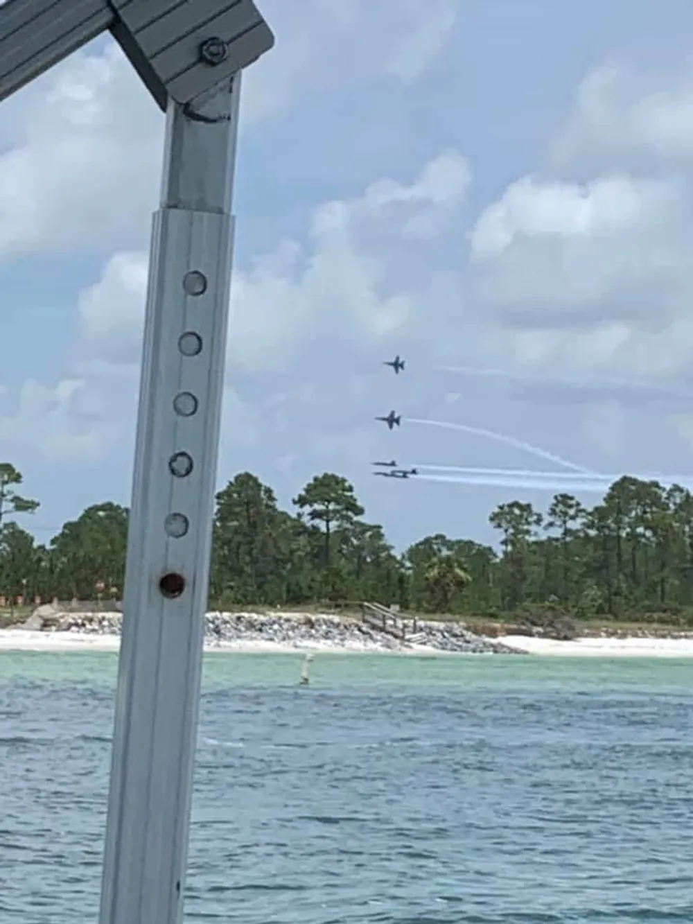 Jets are flying in formation against a cloudy sky over a body of water with a beach and vegetation visible in the background seen from under a structure with a metal beam in the foreground