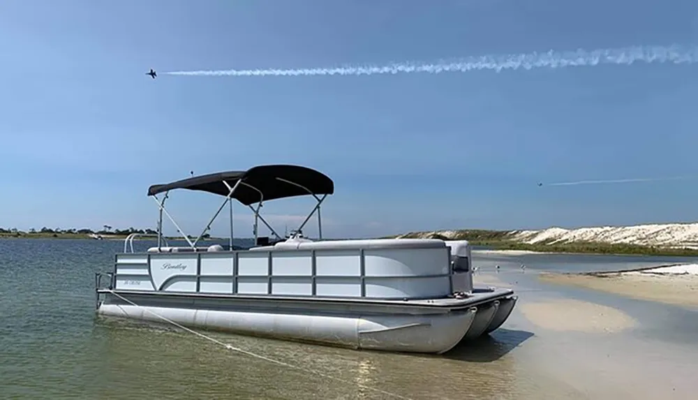 A pontoon boat is beached on a sandy shore with a jet leaving a contrail in the clear blue sky above