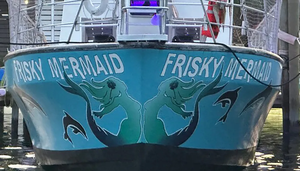 The image shows the bow of a boat named FRISKY MERMAID featuring artwork of two mermaids and marine life painted on its hull