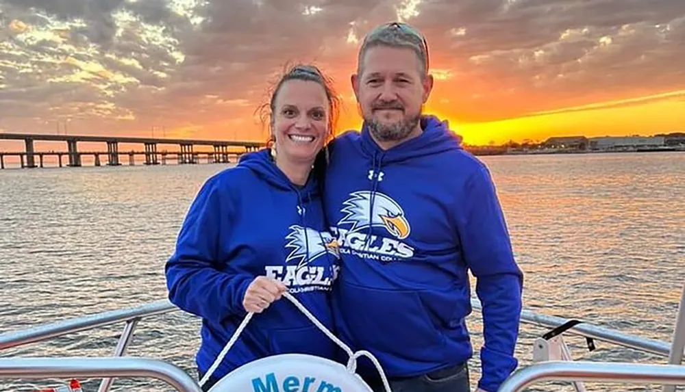Two people are smiling for a photo on a boat with a stunning sunset and a bridge in the background