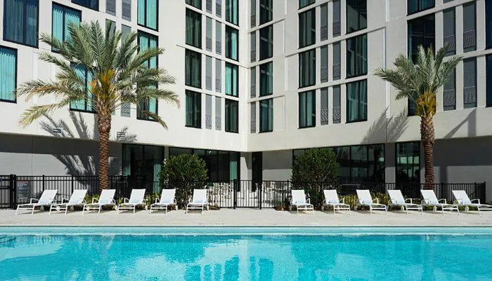 This image shows a row of white lounge chairs lined up beside a swimming pool framed by palm trees and a modern building facade