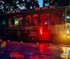 A vibrantly lit theme-decorated trolley bus at night possibly for a haunted tour sits by a curb reflecting its red and yellow lights onto the wet ground