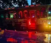 A vibrantly lit theme-decorated trolley bus at night possibly for a haunted tour sits by a curb reflecting its red and yellow lights onto the wet ground