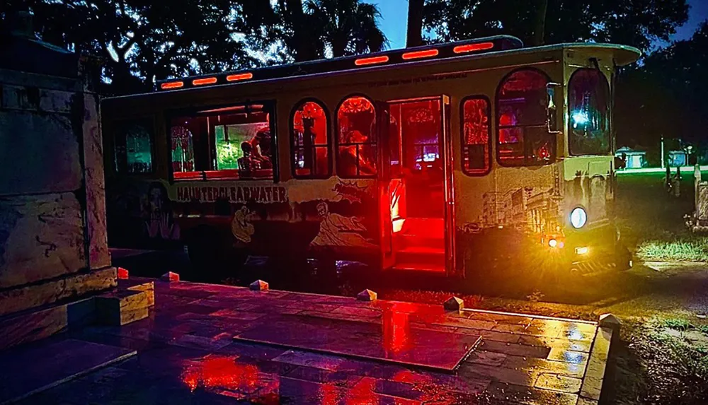 A vibrantly lit theme-decorated trolley bus at night possibly for a haunted tour sits by a curb reflecting its red and yellow lights onto the wet ground