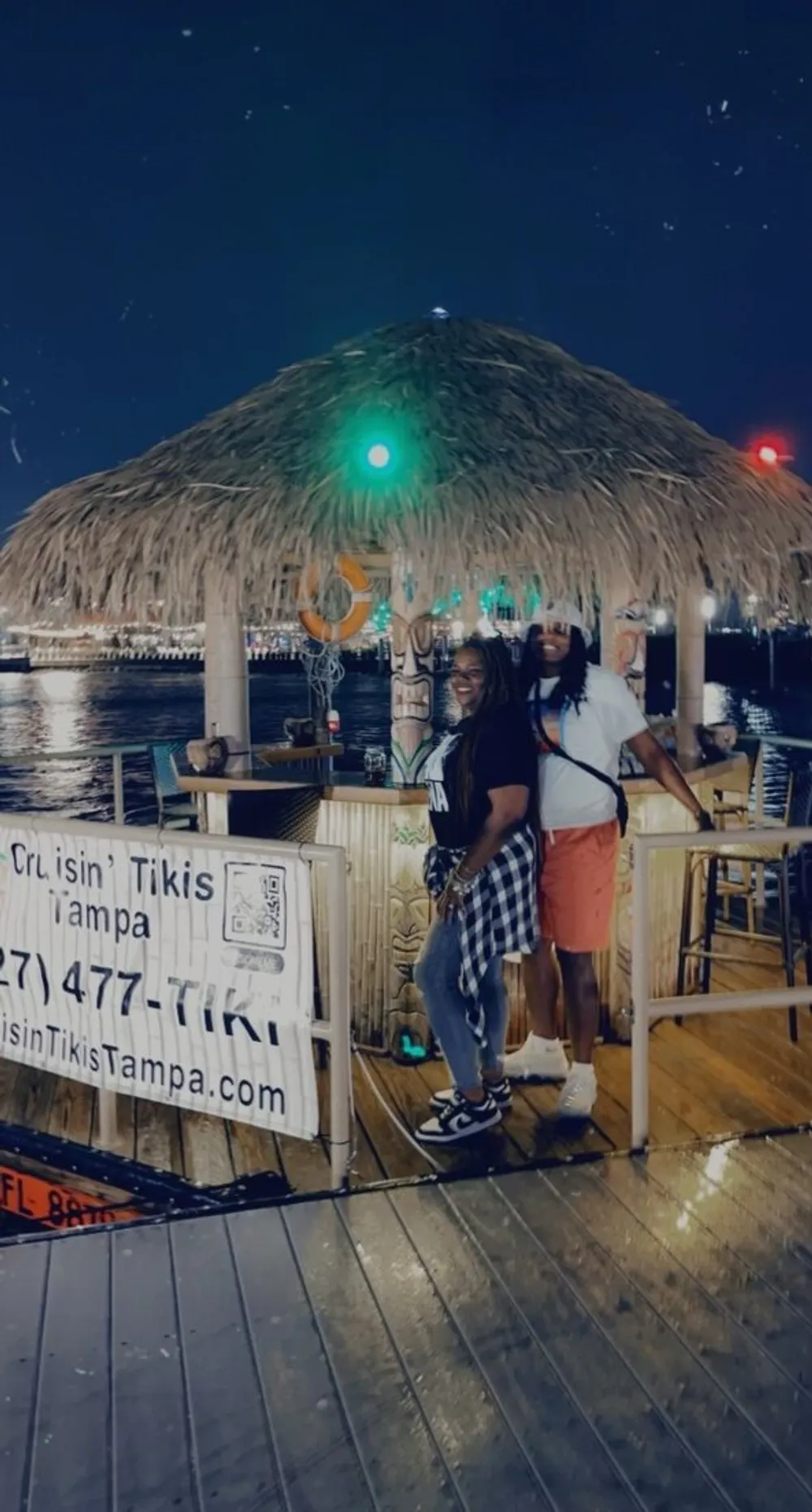 Two people are posing with smiles on a dock next to a thatched-roof structure adorned with festive lights at night giving off a cheerful tropical vibe