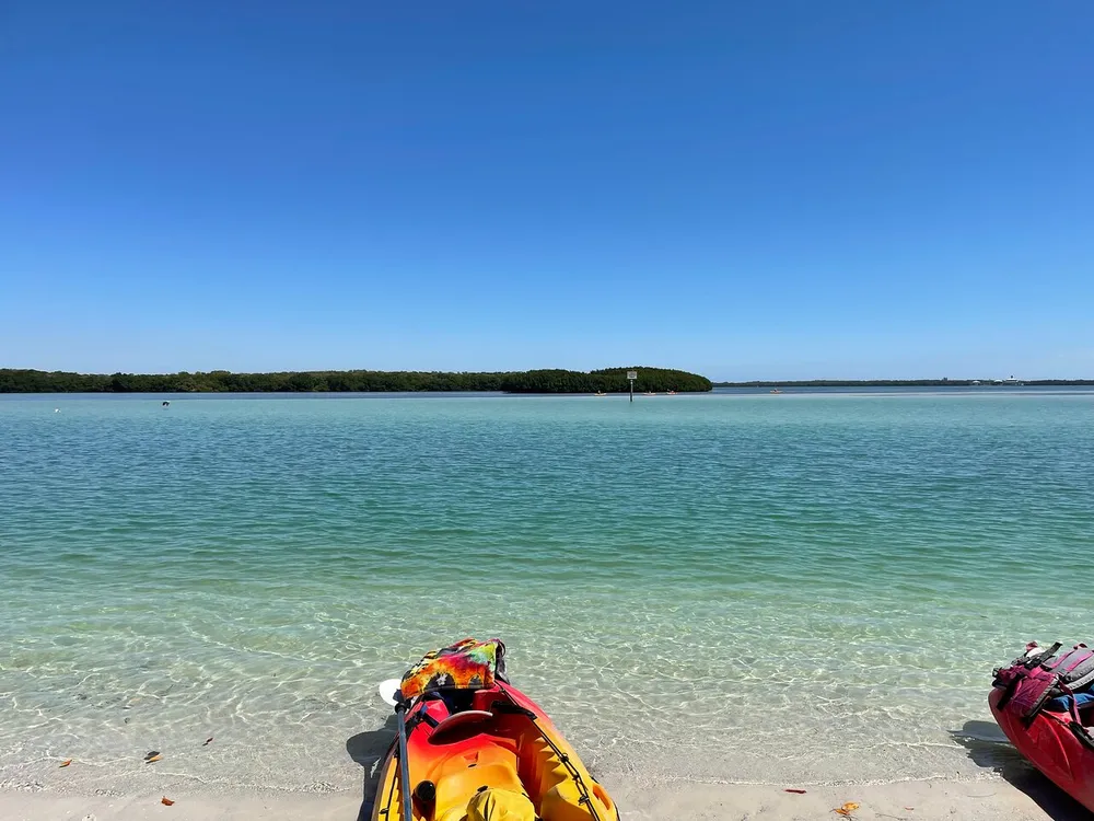 The image shows a sunny day at a clear beach with a tip of a yellow kayak in the foreground and calm turquoise waters leading to a green tree-lined shore in the distance