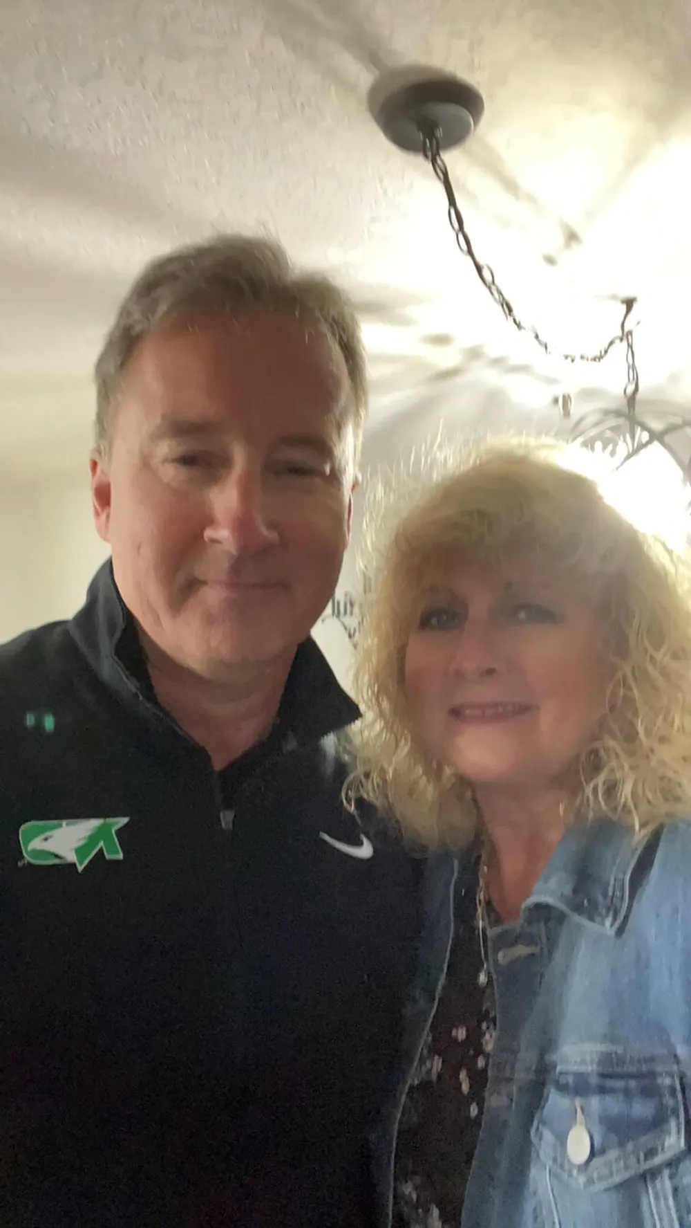 A man and a woman are smiling for a selfie indoors with a light fixture visible in the background