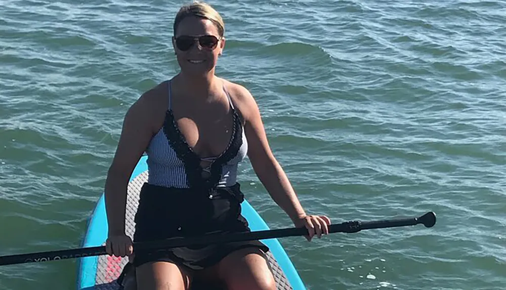 A person is kneeling on a paddleboard in calm waters holding a paddle and smiling towards the camera